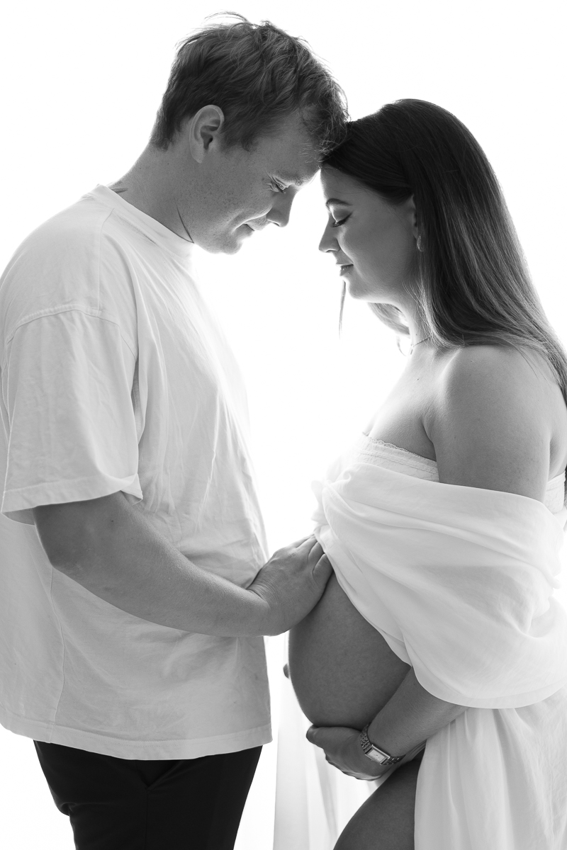 Svartvit parbild på gravid i studiomotljus, panna mot panna och pappans hand på den växande gravidmagen.
