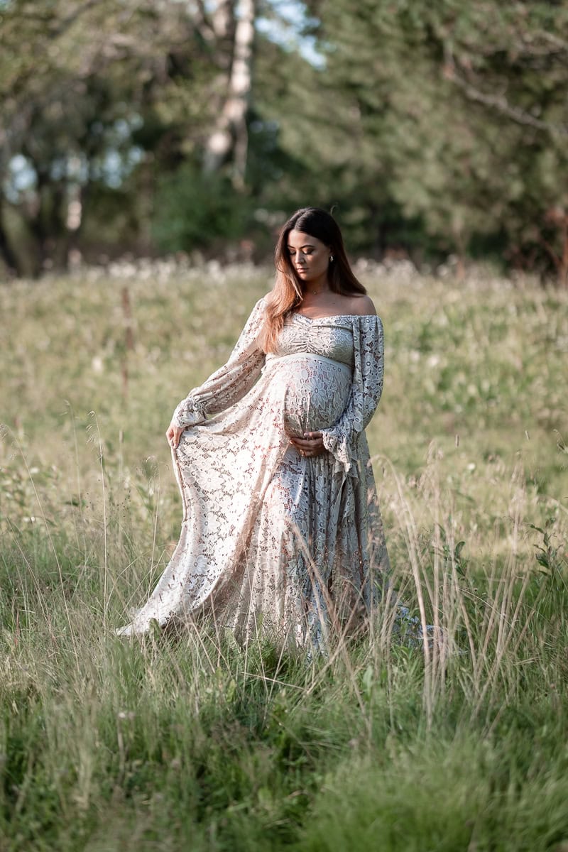 En gravid kvinna klädd i en beige spetsklnning stående mitt på en sommaräng.