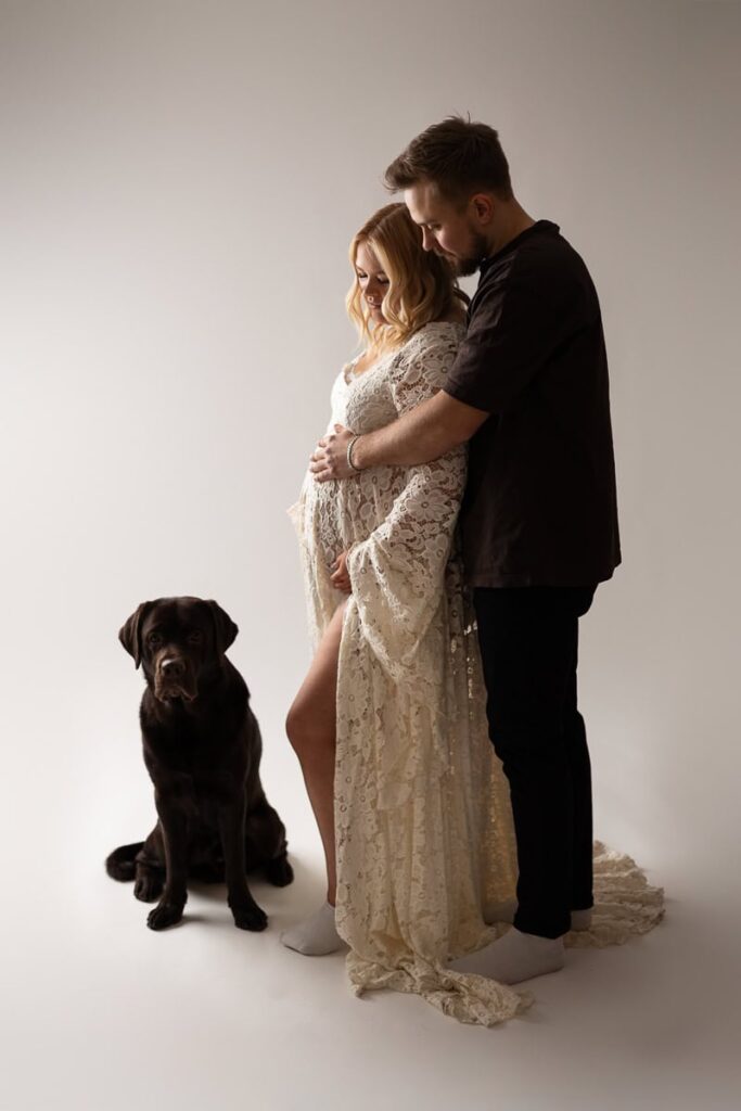 Gravid i lyxig spetsklänning tillsammans med partner och deras hund.